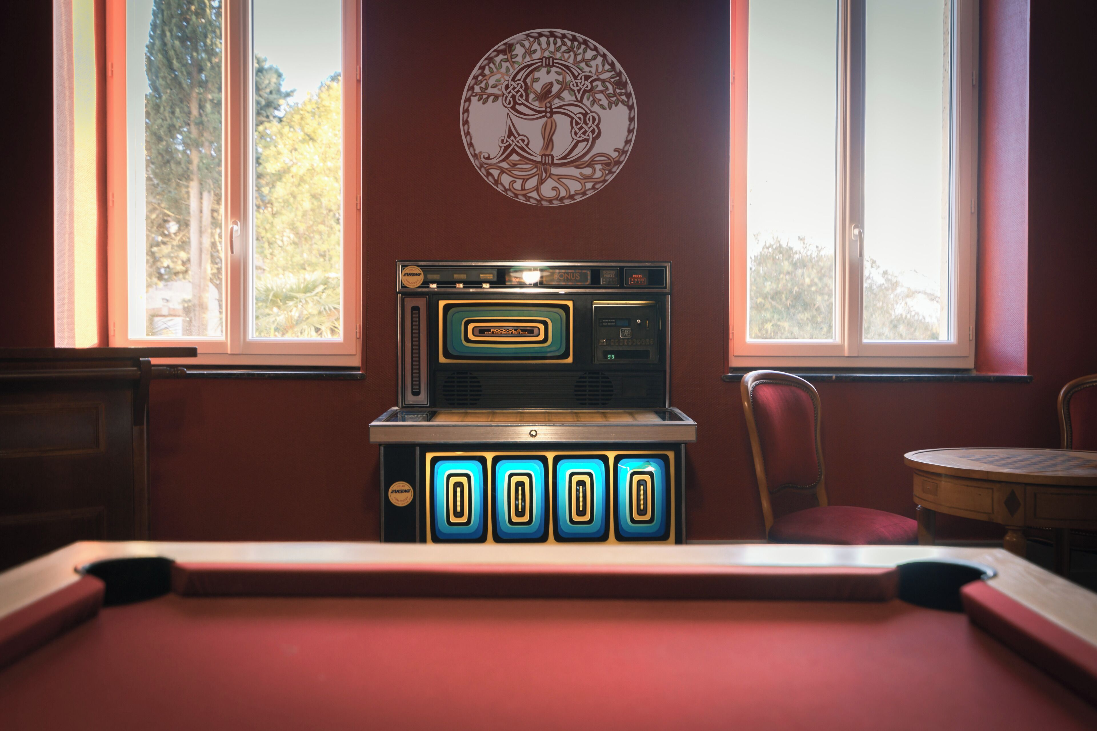 A pool table in a room with a jukebox, a red wall, and a window with a view of trees.