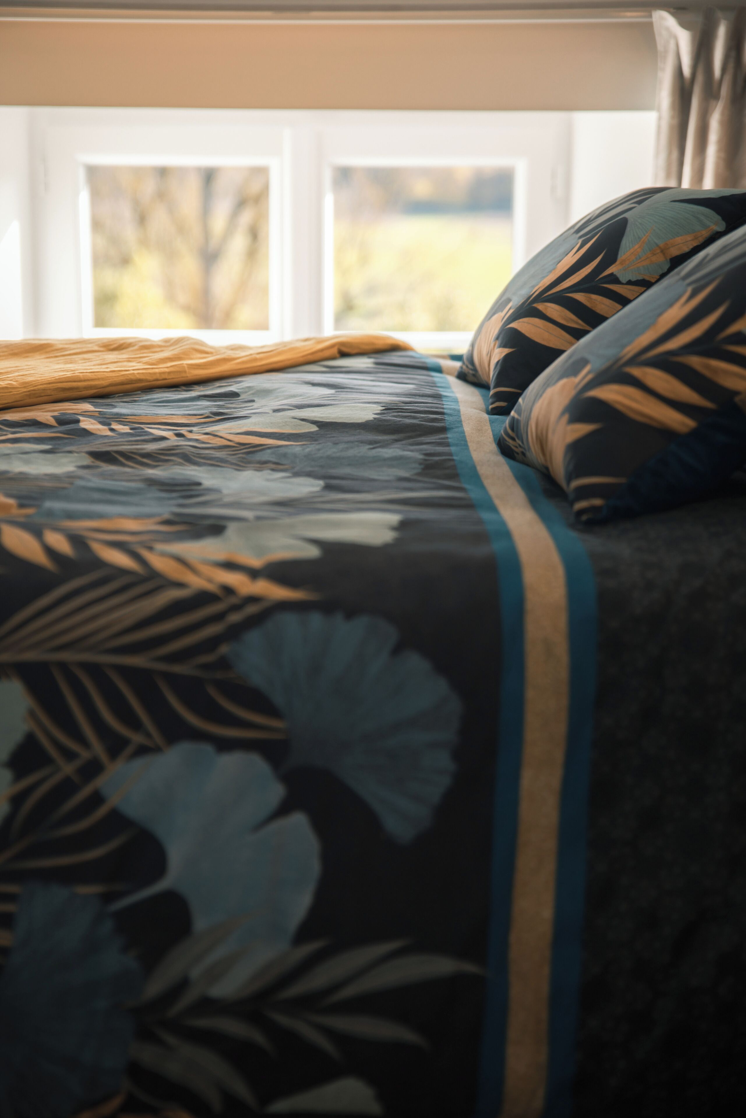 A bed with a floral-patterned bedspread and pillows, a window with curtains, and a view of trees outside.