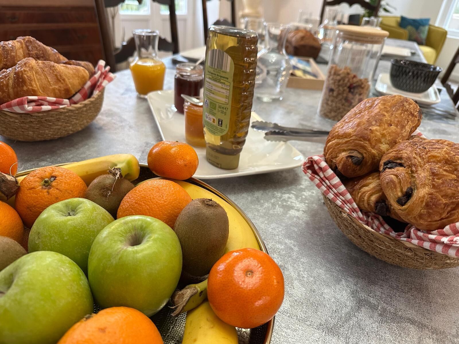 Une table de petit-déjeuner avec des croissants, un panier de fruits, un pot de miel et un verre de jus d’orange.