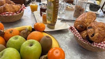 Une table de petit-déjeuner avec des croissants, un panier de fruits, un pot de miel et un verre de jus d’orange.