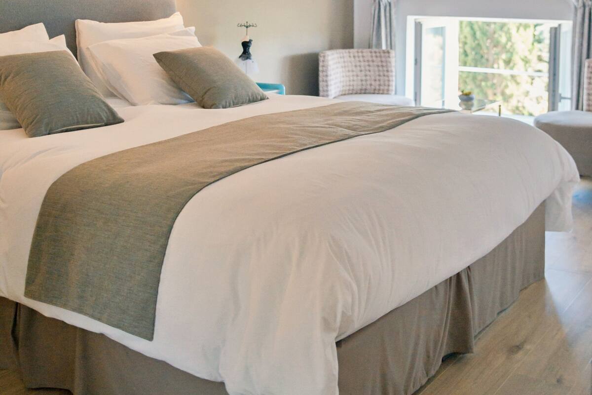 A large bed with a grey and white duvet, a bedside table with a lamp, and a window with curtains.