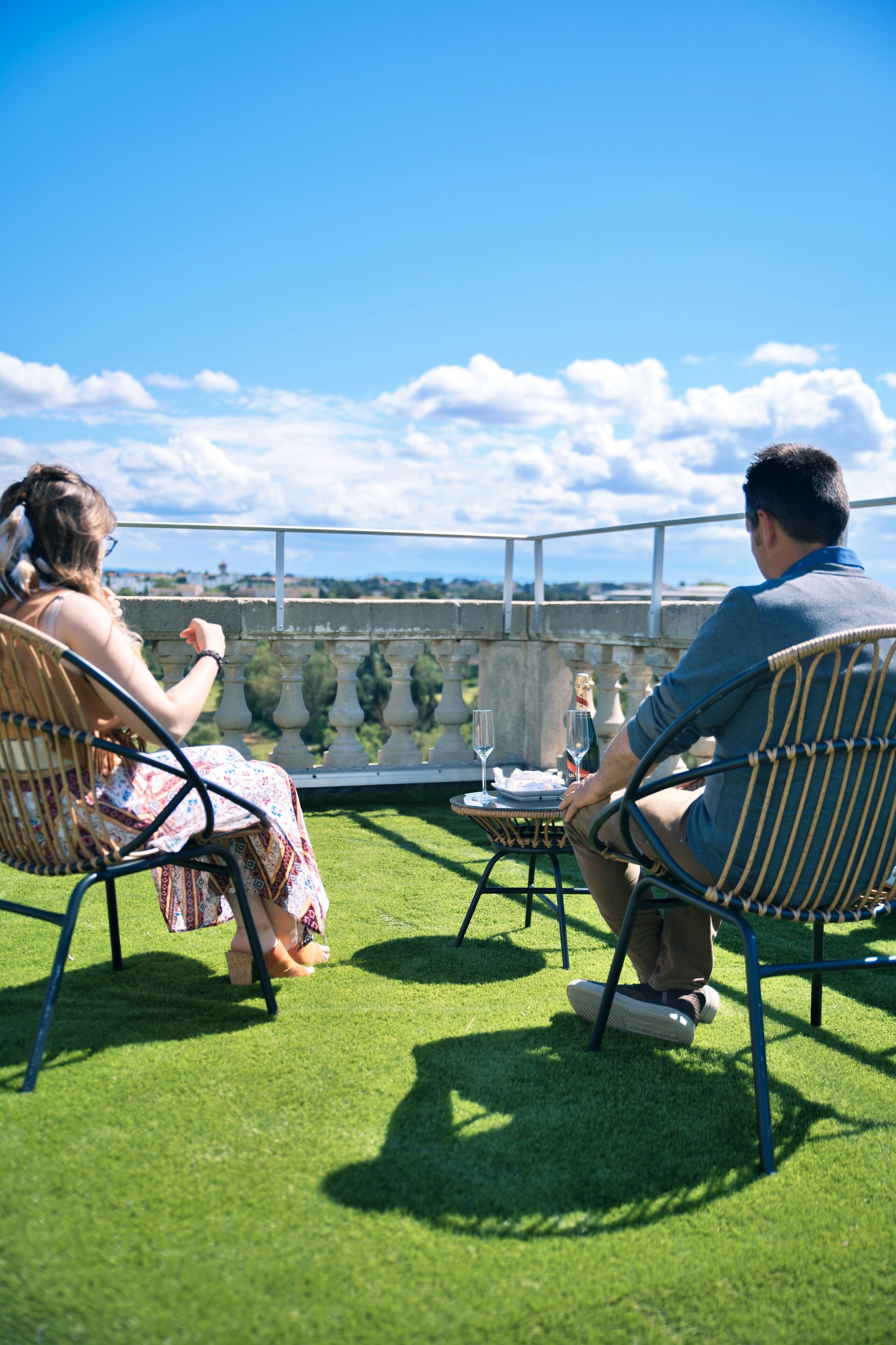 Two people seated on wicker chairs facing a table with a bottle and glasses, on a balcony with a view of a stone balustrade and greenery.