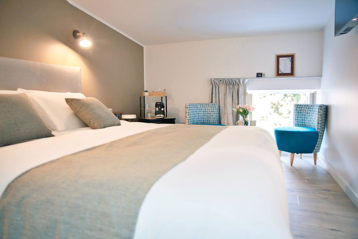 A modern hotel room with a large bed, a nightstand with a coffee maker, a chair, and a view of the outdoors.