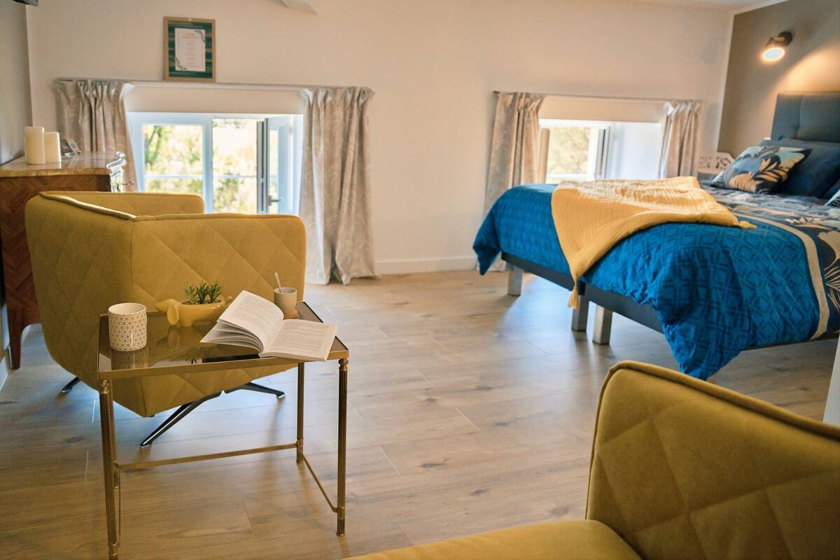 A modern bedroom with a bed, a reading chair, a small table with a book, and a window with curtains.