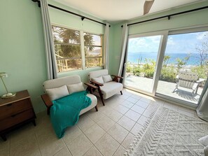 A room with two white sofas, a teal throw blanket, a wooden nightstand, and a view of the ocean through large windows.