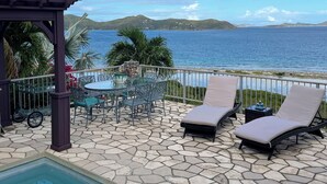 A poolside area with lounge chairs, a table, and chairs, overlooking a body of water and distant hills. - Thalassa. A Seaside Retreat with Pool & Infinity Oceanfront Sunrise Views (St. John)