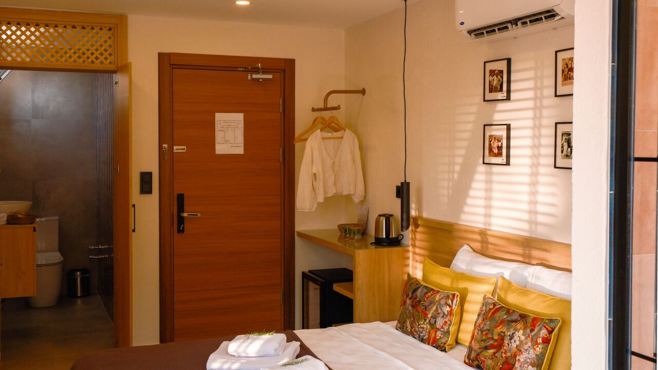 Habitación de hotel con puerta de madera, cama con almohadas estampadas, mesita con hervidor y aire acondicionado instalado en la pared.