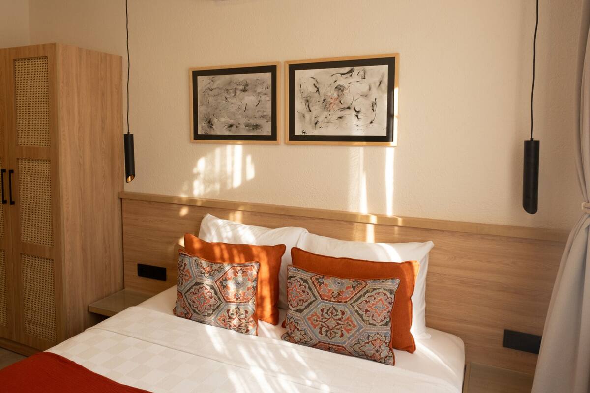 A hotel room with a wooden headboard, a bed with white bedding, two patterned pillows, and two abstract paintings on the wall.