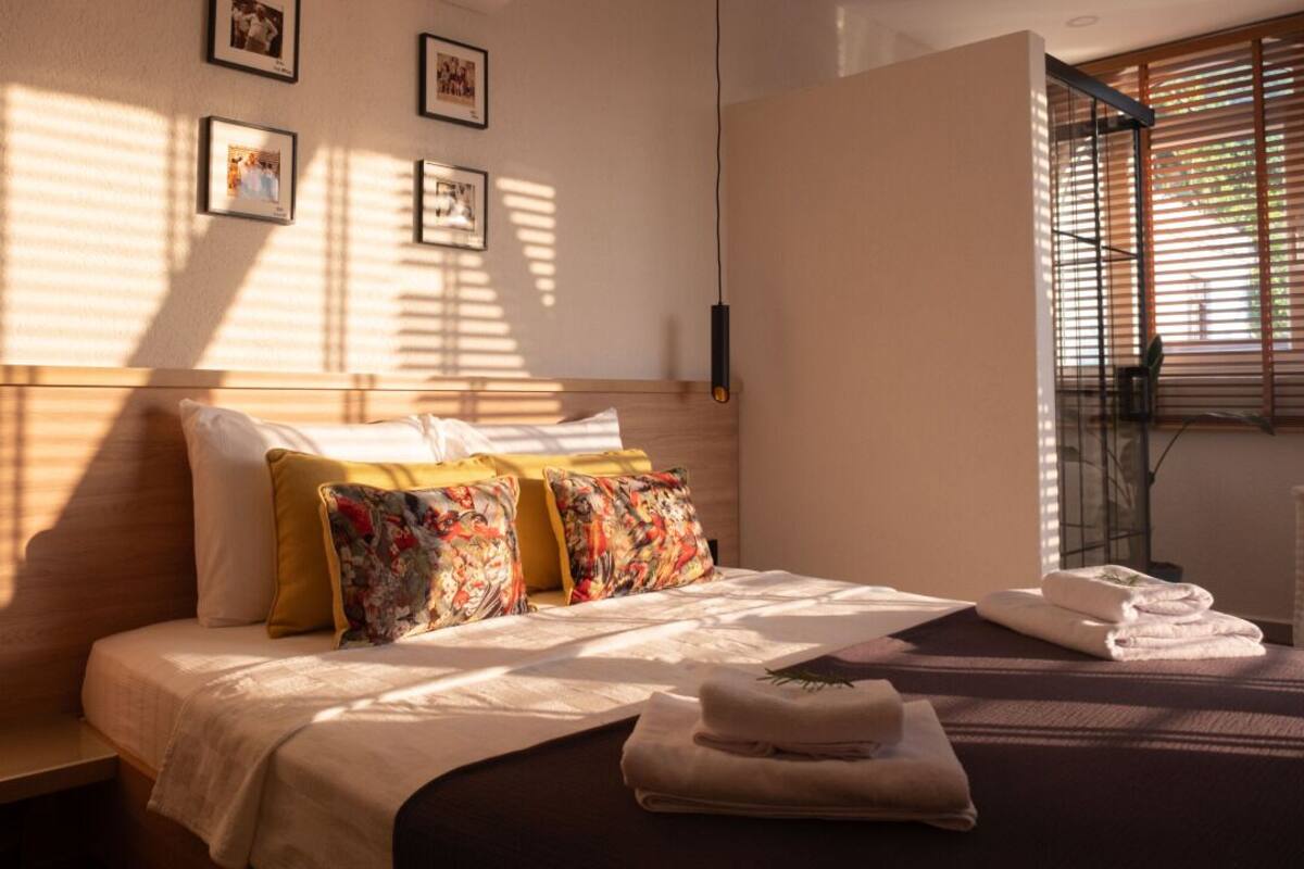 A neatly made bed with floral pillows, a bedside table with a towel, and a window with blinds.