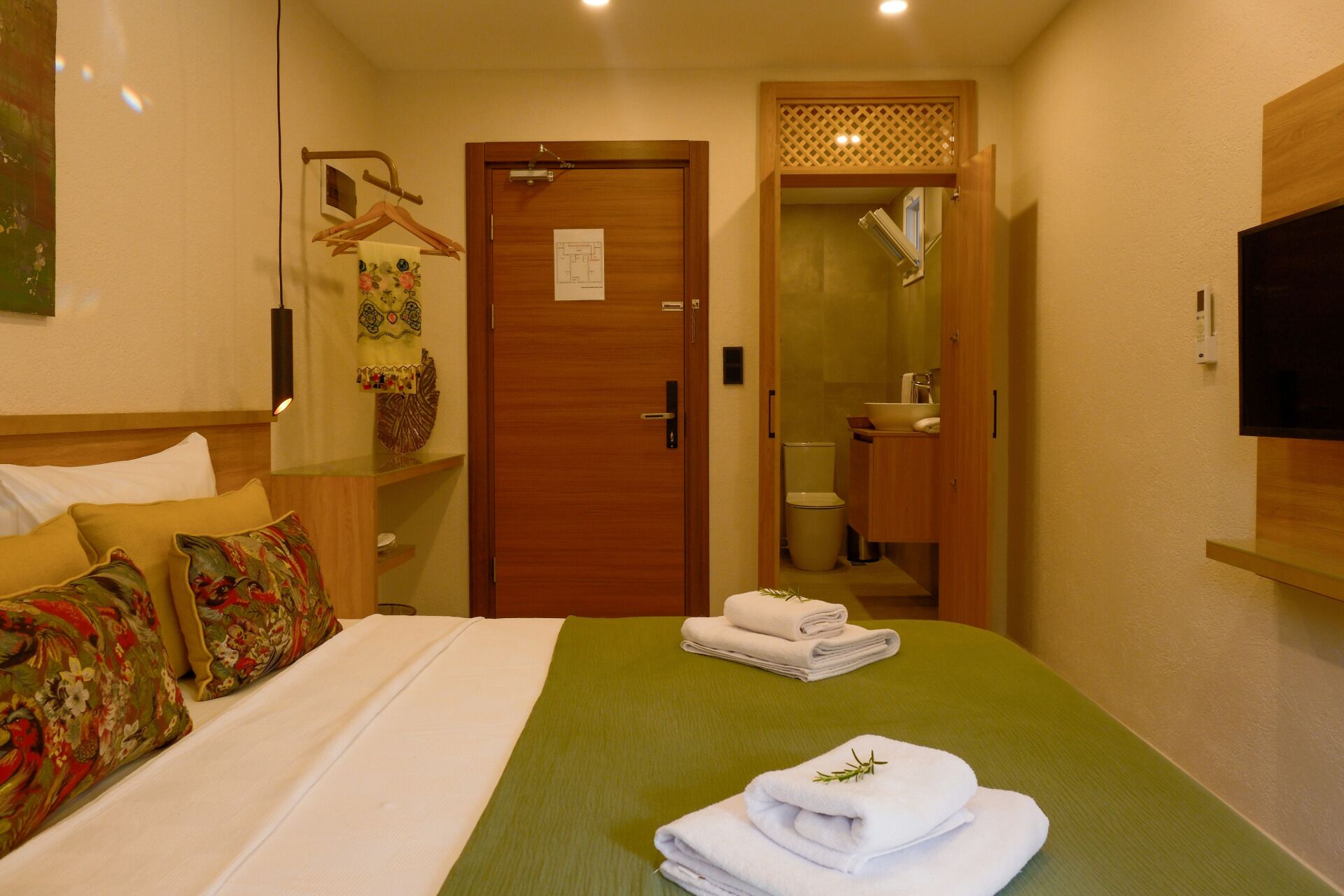 Een hotelkamer met een bed, een groene plaid, een houten deur, een badkamer met toilet en wastafel, en een aan de muur bevestigde televisie.