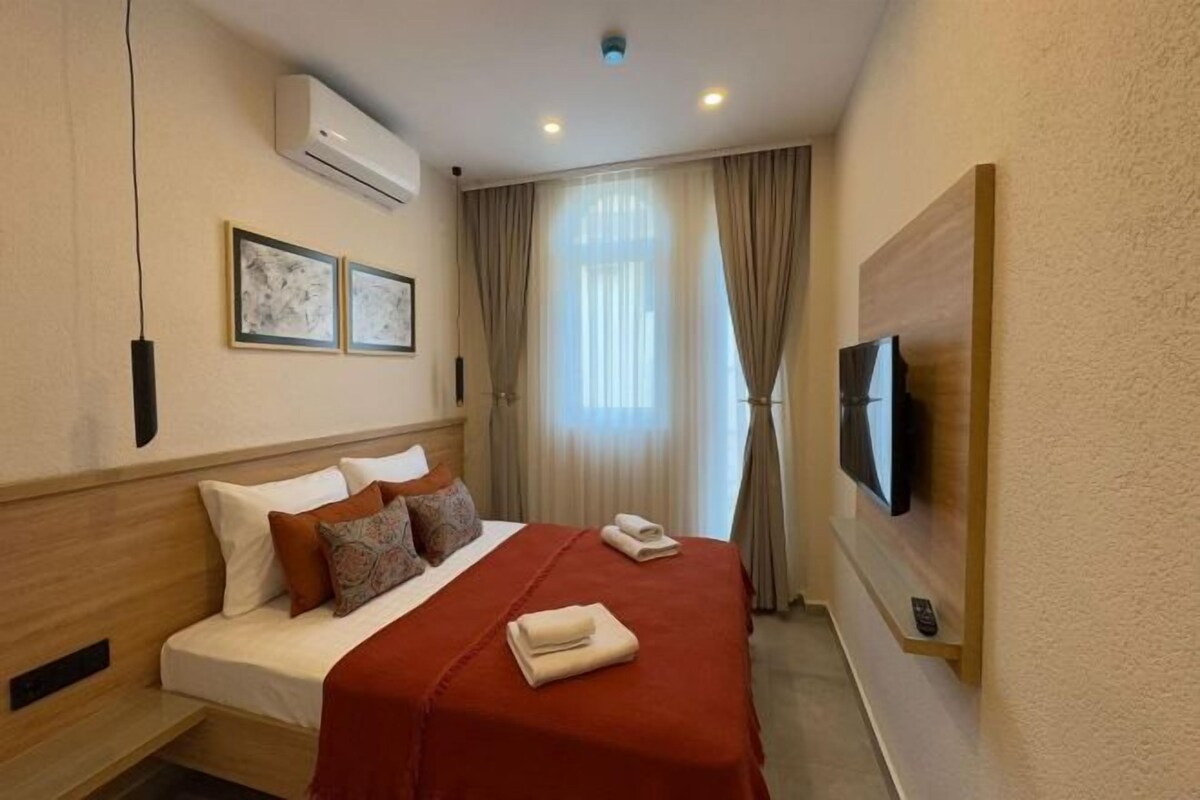 A hotel room with a bed, air conditioning unit, curtains, a television, and framed artwork on the wall.