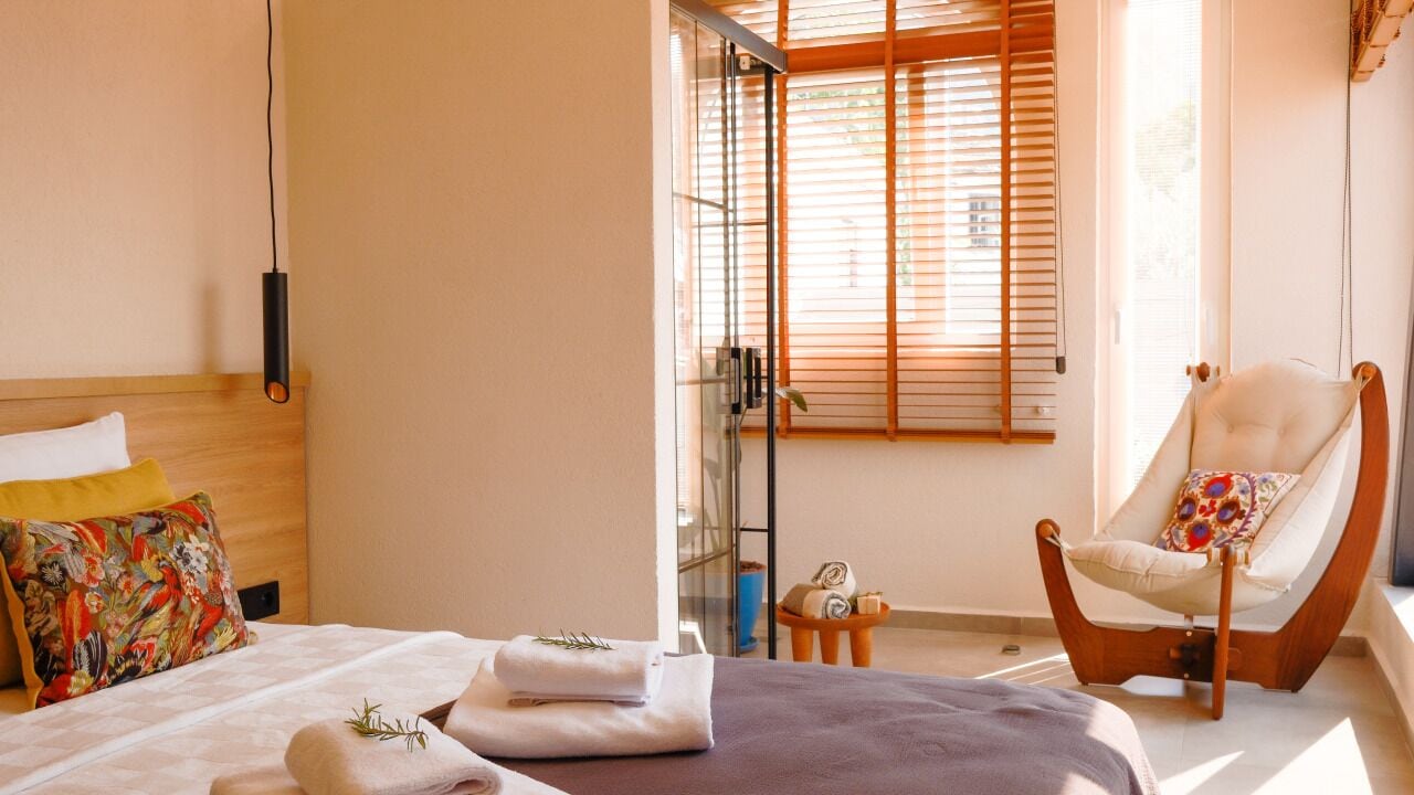 A hotel room with a bed, a chair, a small table, and a window with blinds.