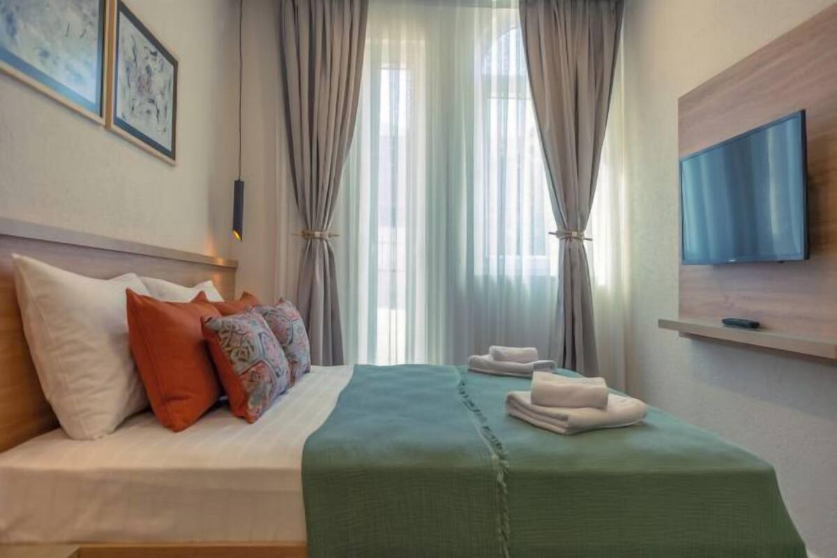 A hotel room with a bed, pillows, a green bedspread, a TV on the wall, and curtains.