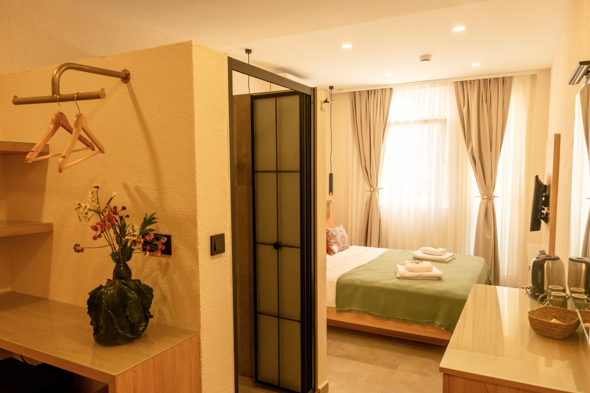 A hotel room with a bed, a small table, a chair, a wall-mounted shelf with a vase of flowers, and a door leading to another room.