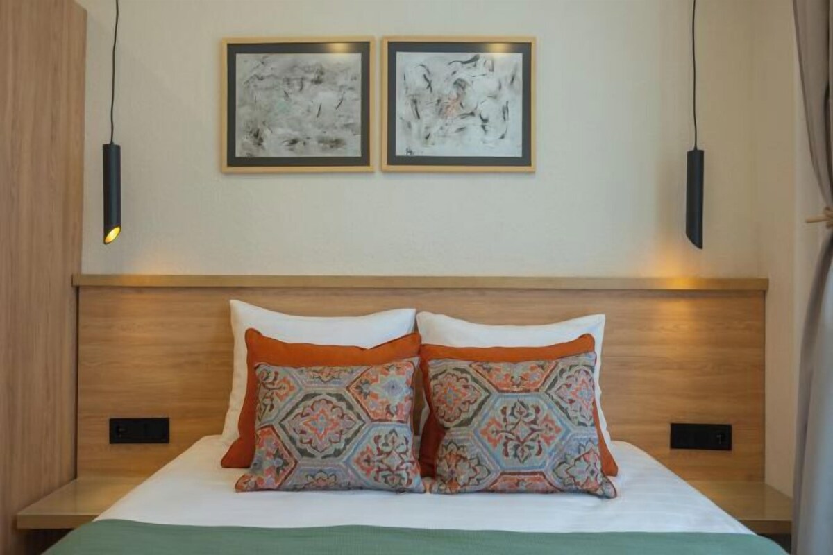 A hotel room with a wooden headboard, two framed abstract paintings, a bed with patterned pillows, and bedside lamps.