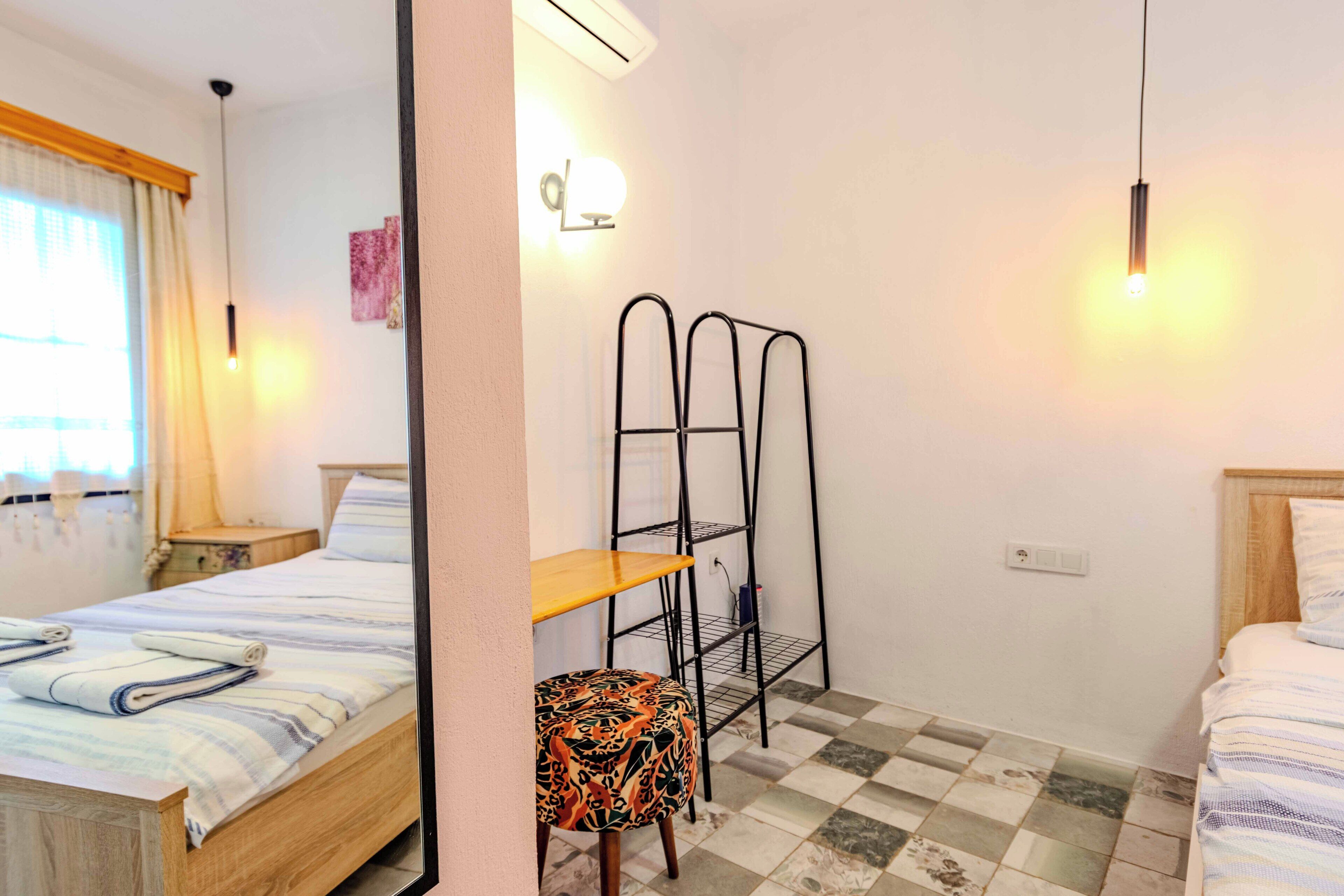 Une chambre avec deux lits, une table de chevet en bois, une petite table, une chaise, une étagère fixée au mur et un sol à carreaux.