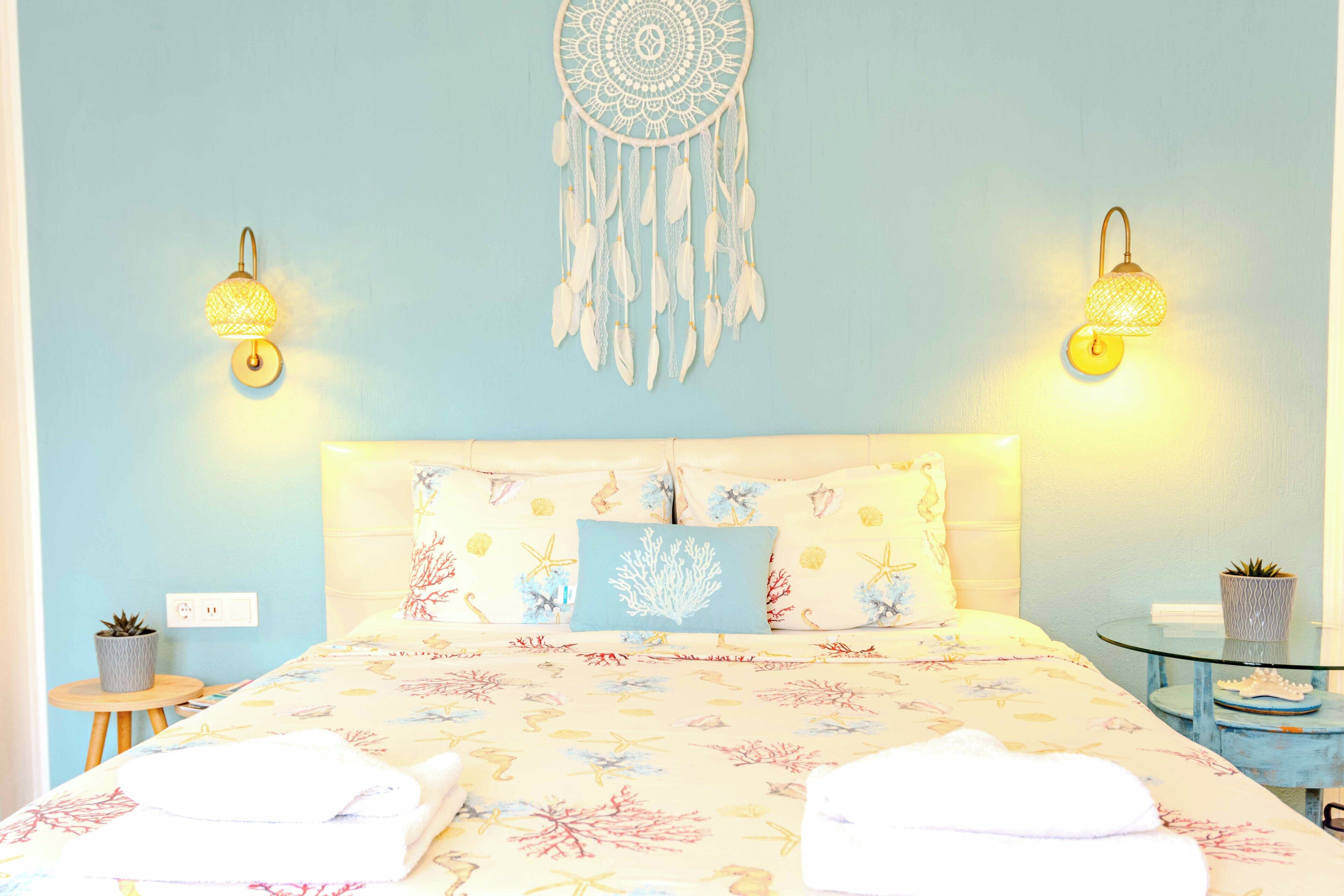 A bed with a floral bedspread, two wall-mounted lamps, a dreamcatcher, and a small table with a plant.