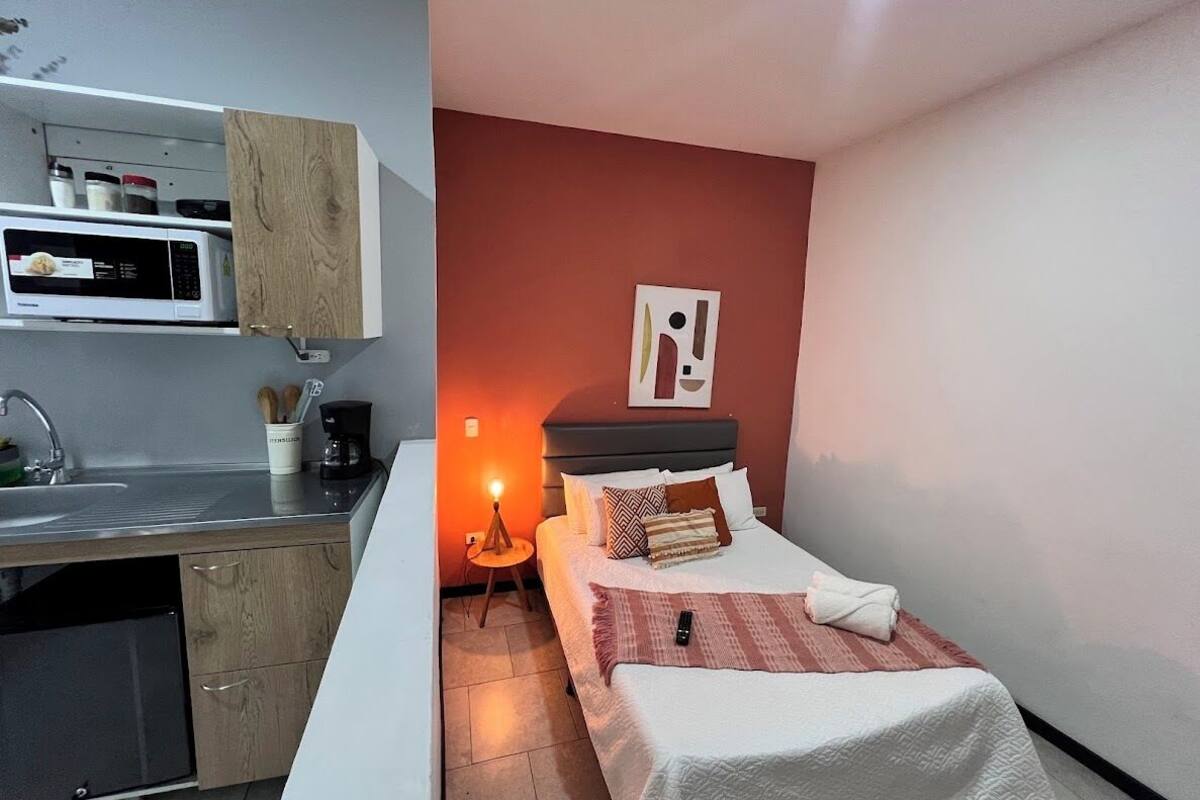 A compact hotel room with a kitchenette, a bed with a red wall, and a bedside lamp.