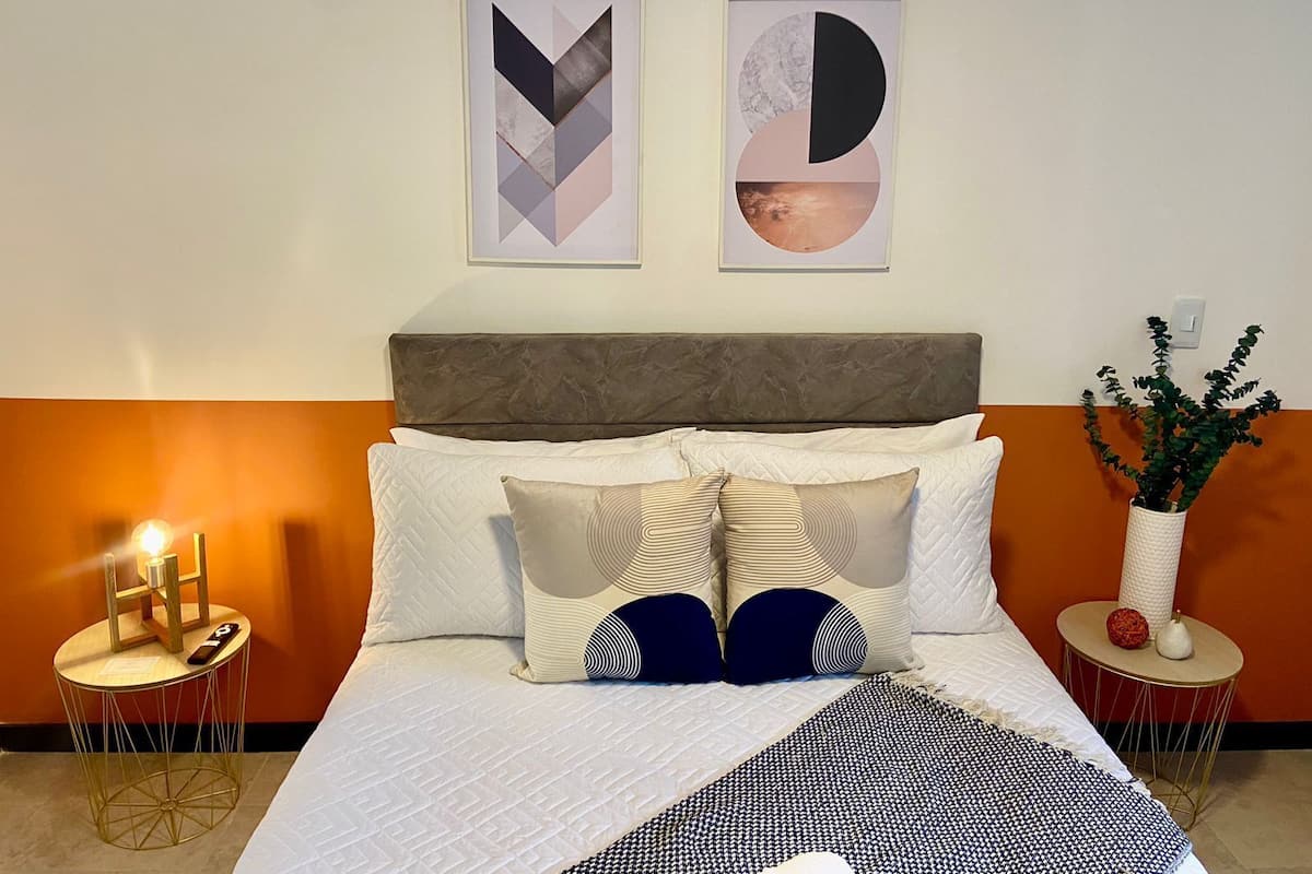 A neatly made bed with a white duvet, multiple pillows, and a black and white throw blanket. There are two framed abstract artworks above the bed, a small table with a candle and a decorative item, and a potted plant on a side table.