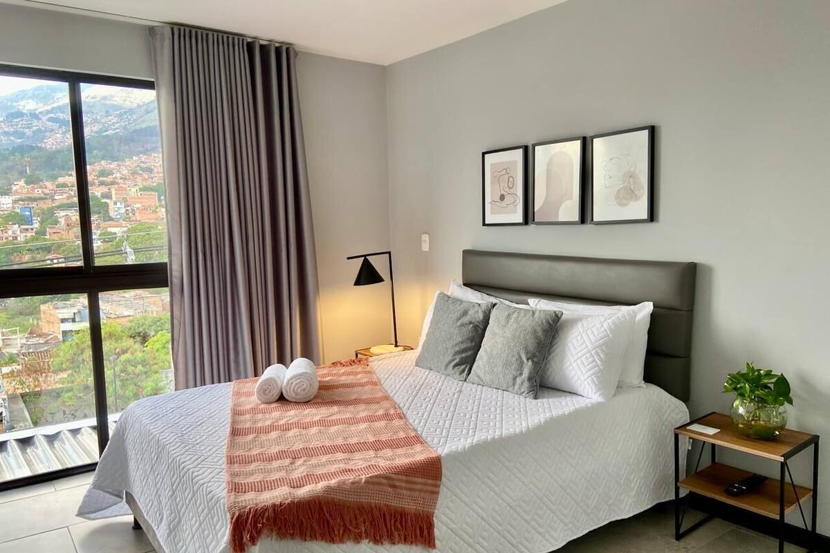 A modern hotel room with a large bed, a view of a cityscape, and a small side table with a plant.