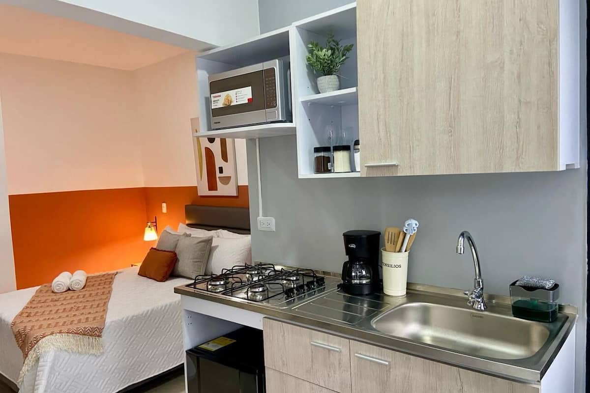 A compact hotel room with a kitchenette, a bed with pillows, a bedside lamp, and a small potted plant.