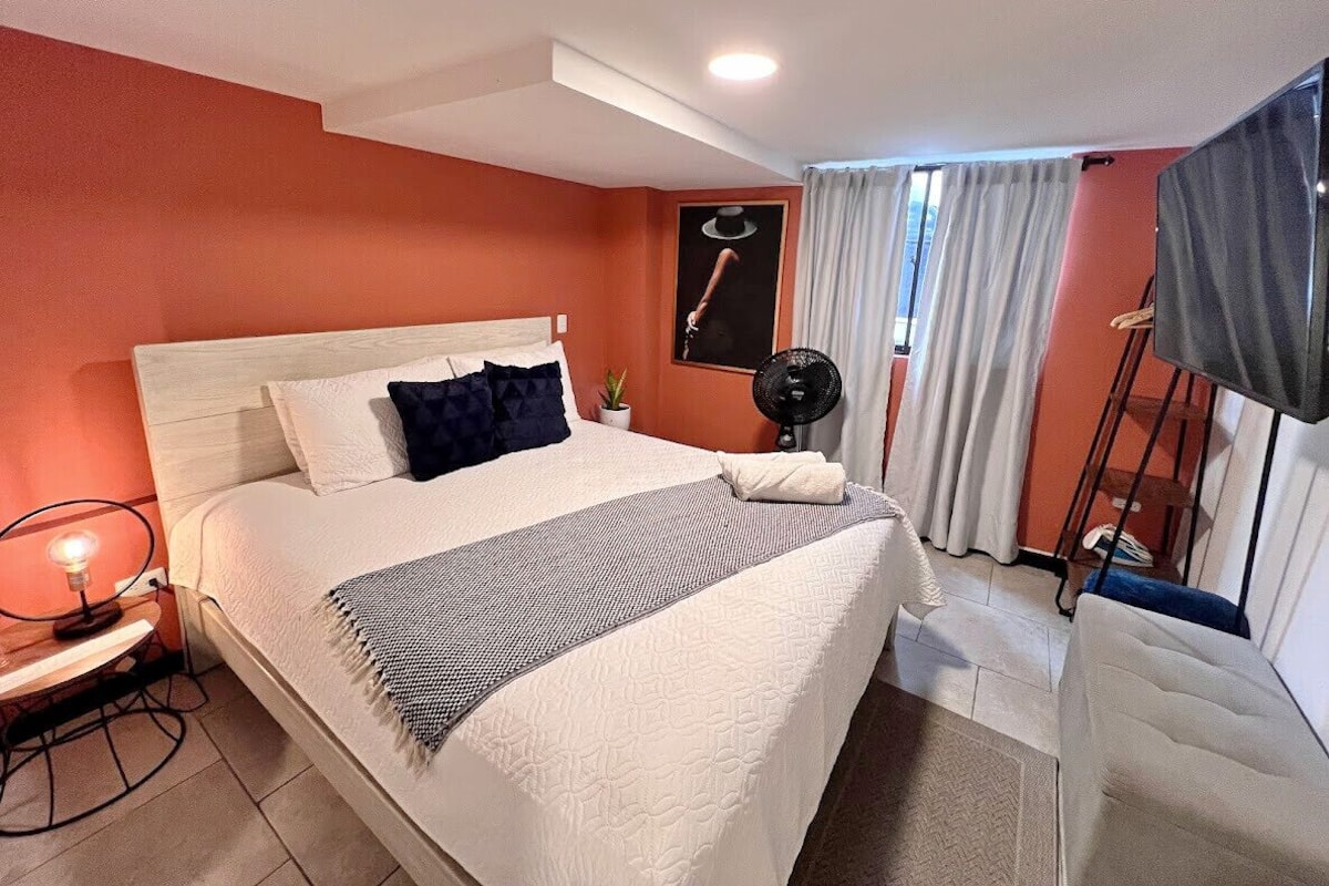 A hotel room with a large bed, a desk with a lamp, a fan, and a TV.