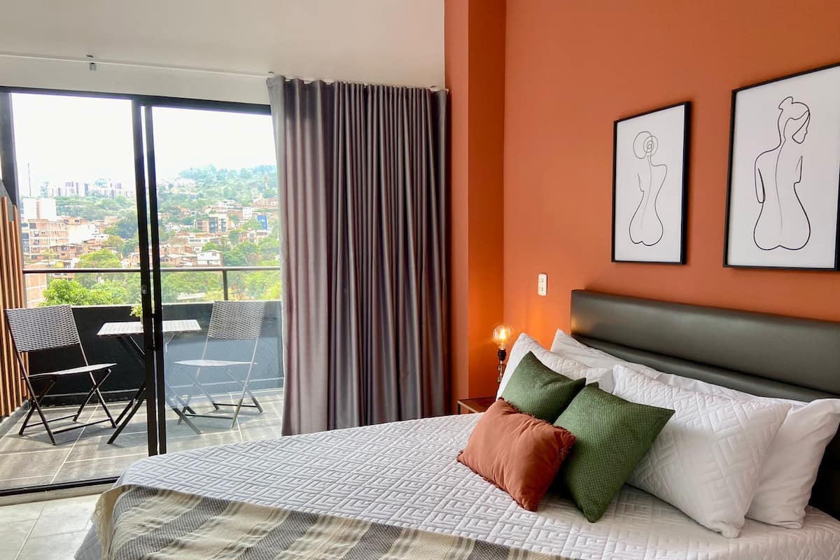 A hotel room with a bed, a balcony with a chair, and two framed artworks on the wall.
