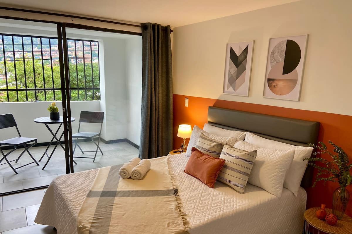 A hotel room with a bed, a nightstand, a lamp, a small table with a vase, and a view of greenery outside.