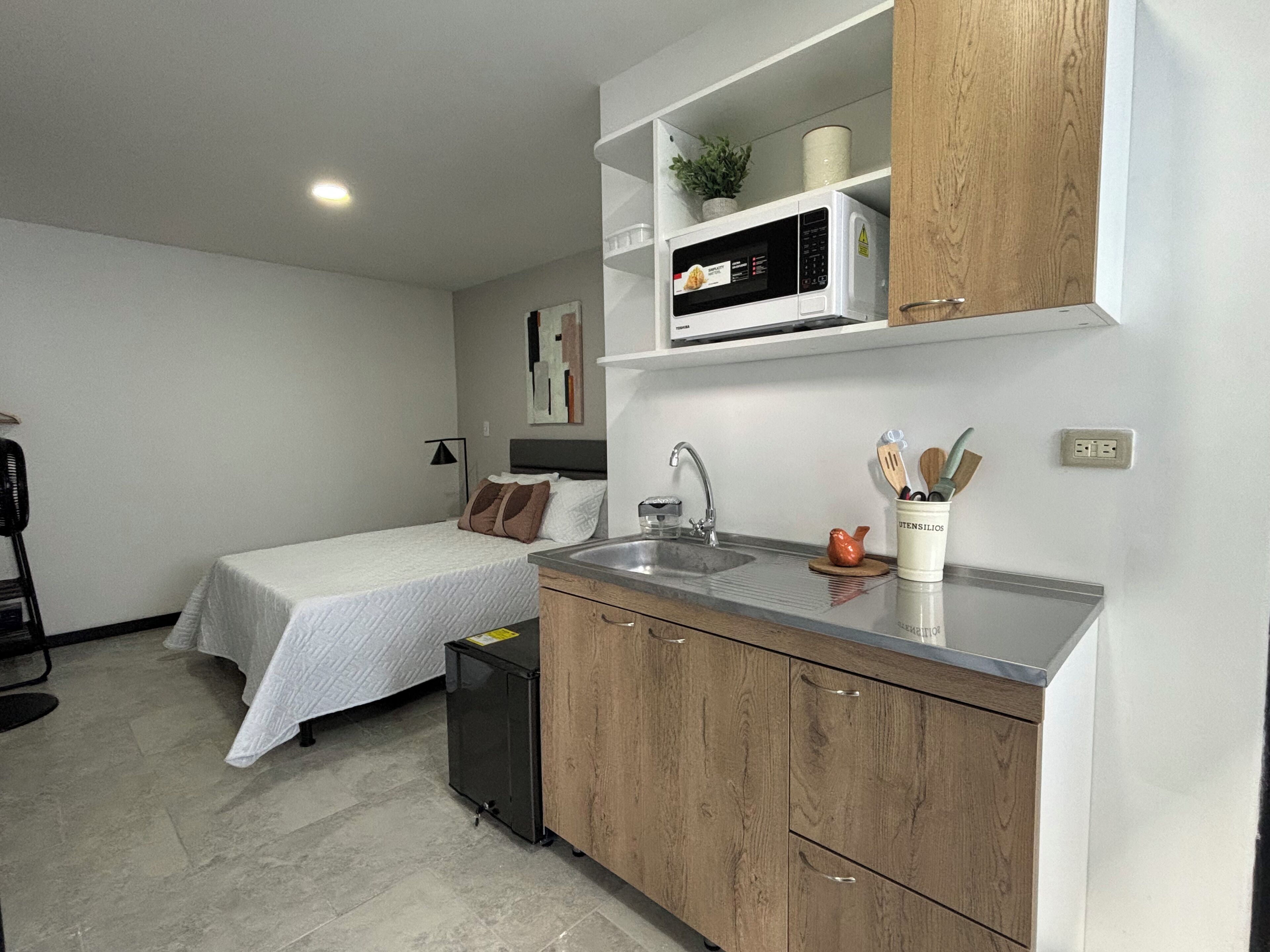 A compact hotel room with a kitchenette, a bed with a white duvet, a small bedside table, and a wall-mounted shelf with a microwave and a potted plant.