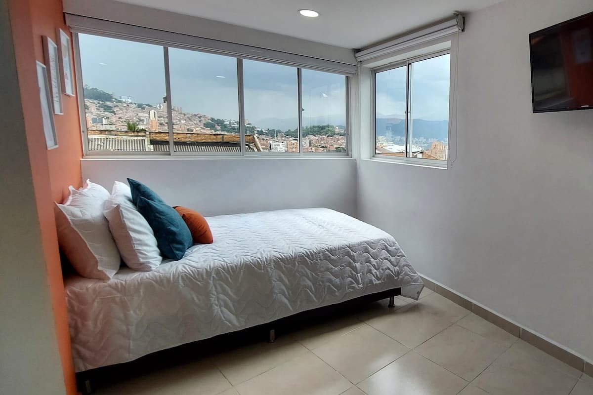 A bedroom with a bed, a TV mounted on the wall, and a city view through large windows.