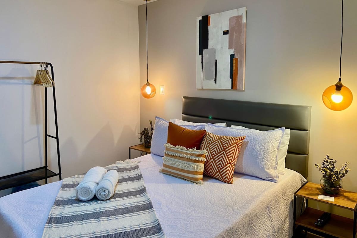 A neatly made bed with a striped blanket, two pillows, and rolled towels. There is a bedside table with a vase of flowers and a remote control.