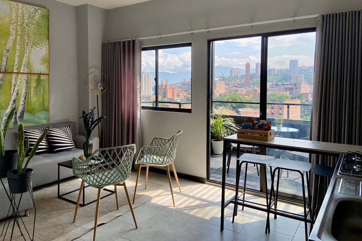 A modern living room with a city view, a dining area with stools, and a kitchen with a sink.