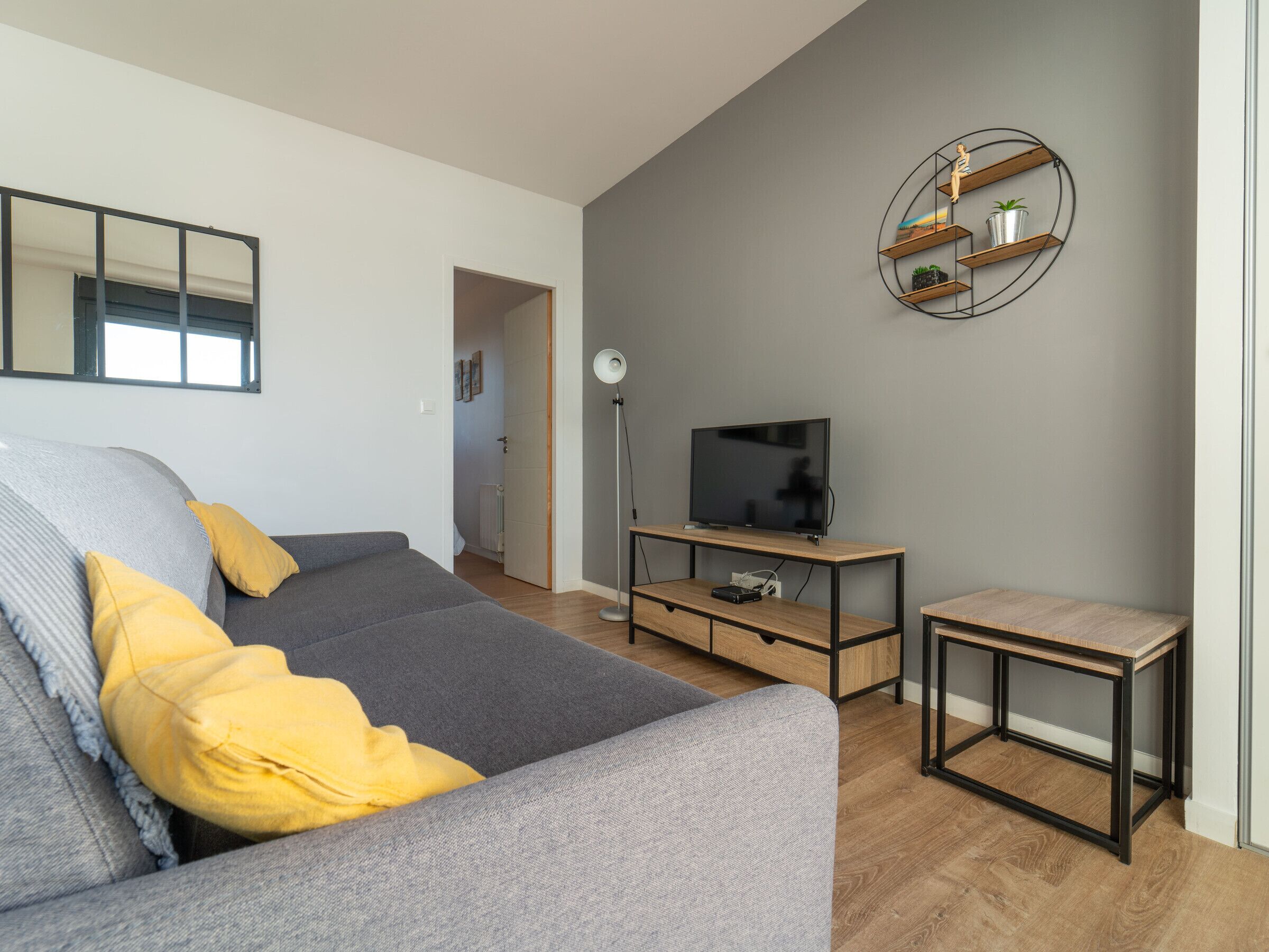 Una sala de estar moderna con un sofá gris, un televisor sobre un mueble de madera, una lámpara de pie y una mesita.