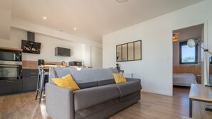 A modern living space with a grey sofa, wooden flooring, a kitchen area with a microwave, stove, and coffee maker, and a bed with a wooden headboard. - Charming apartment 200m from ocean with terrace and parking (Mimizan-Mimizan Plage)