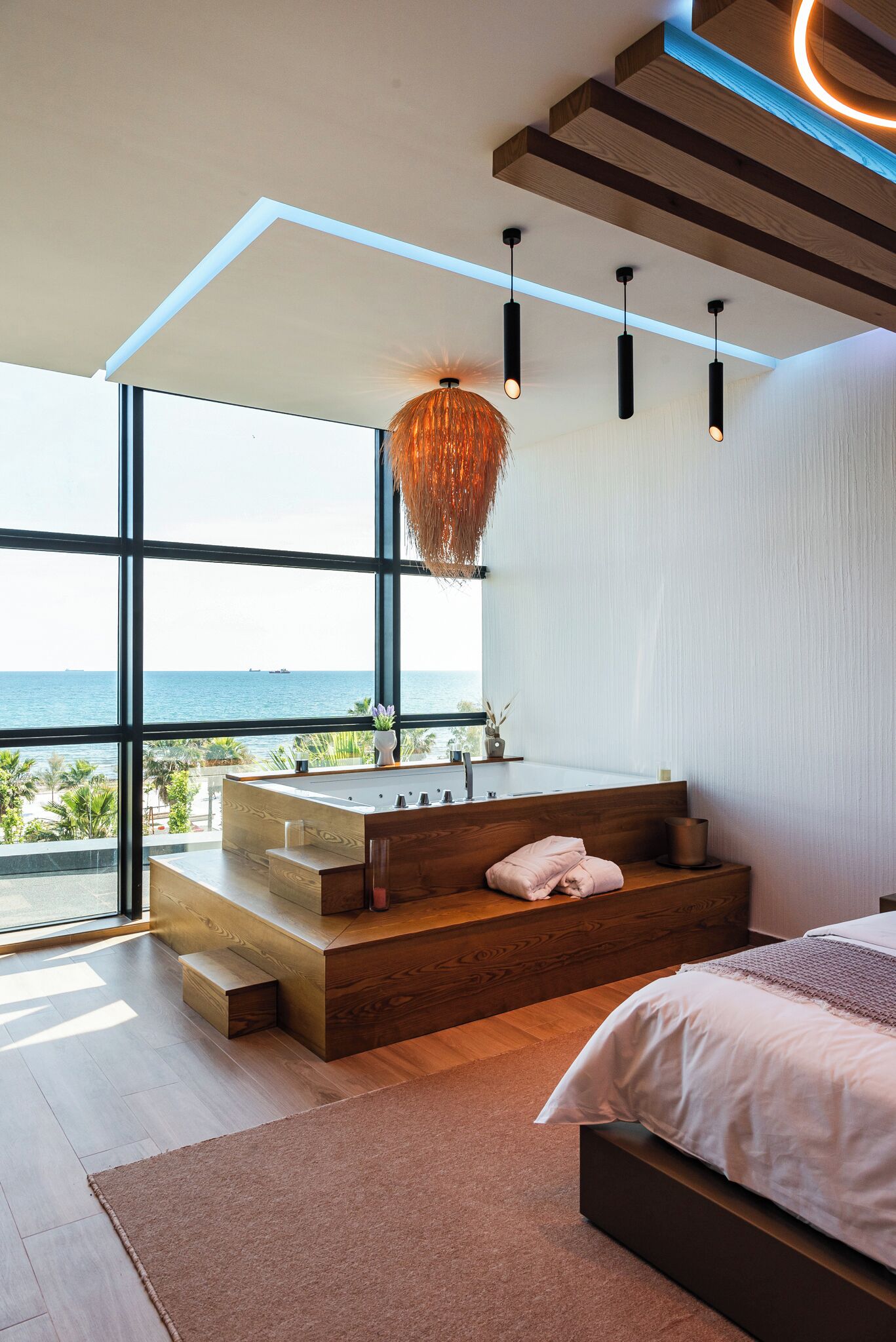 A modern bedroom with a large window, a wooden bench, a bed, and a hanging light fixture.