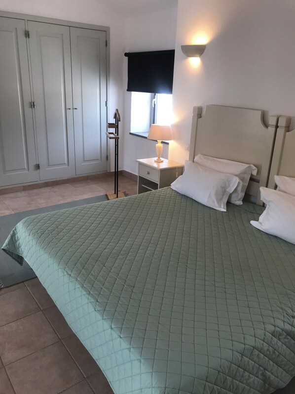 A bedroom with a green quilted bedspread, a bedside lamp, a nightstand, and a wardrobe.