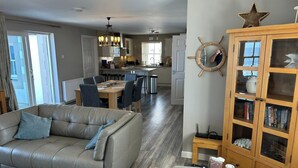 A modern living room with a leather sofa, a dining area with a table and chairs, a kitchen in the background, and a wooden cabinet with books. - Findochty Beach House - Private Garden-beachfront (Buckie)
