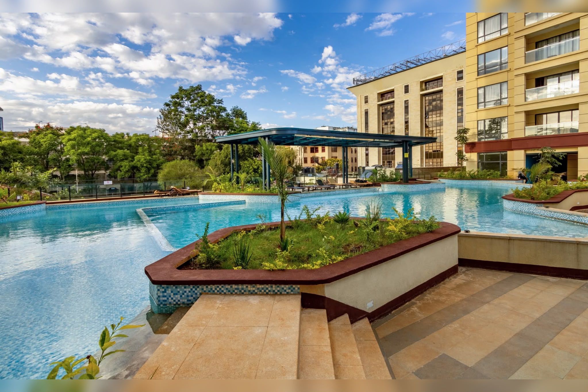 A swimming pool with a covered area, surrounded by a building with multiple balconies and a garden.