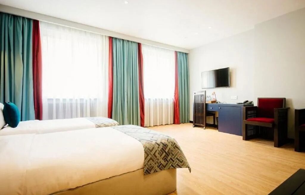 Habitació d'hotel amb dos llits, una taula de treball, una cadira, televisió i finestres grans amb cortines.