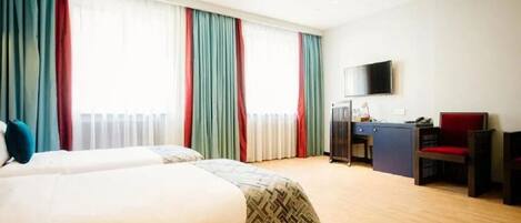 Habitació d'hotel amb dos llits, una taula de treball, una cadira, televisió i finestres grans amb cortines.