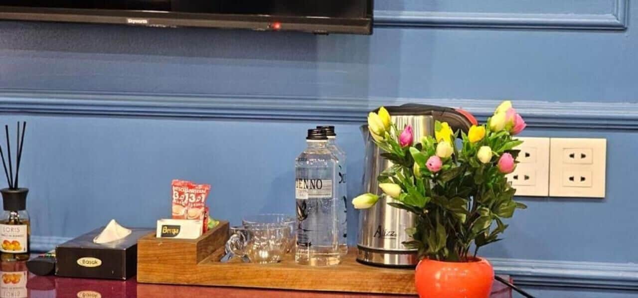 A bar area with a minibar, a bottle of water, a glass, a bottle of alcohol, a pack of tissues, a box of tissues, a vase with flowers, and a candle.