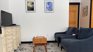 Ein Wohnzimmer mit grauem Sofa, einer Holzkommode und einem kleinen Holzschemel. An der Wand befinden sich zwei gerahmte Bilder.