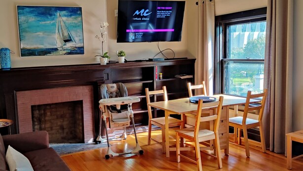 A living room with a fireplace, a television mounted on the wall, a dining table with chairs, and a large window with a view of the outdoors. - Historic 3-Bedroom Guest Suite · Prime Forest Park Location in Central West End (St. Louis)