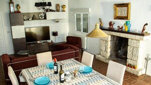 Zona pranzo con tovaglia fantasia, piatti blu e bottiglie di vino. Un televisore è montato sopra un divano scuro. Si intravede uno sfondo con un caminetto e oggetti decorativi.