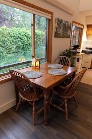 A dining area with a wooden table and chairs, a view of greenery outside, and a kitchen in the background. - Buena Vista Garden Studio Apartment: STR-043 (Buena Vista)