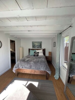 Un dormitorio con cama, un cuadro sobre la cama, una ventana con cortinas y suelo de madera.