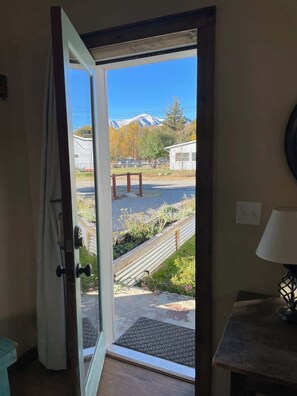 A room with a door leading to a view of a mountain, a small outdoor seating area, and a lamp on a table.