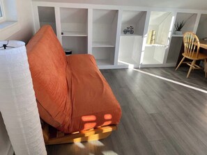Ein Zimmer mit einer orangefarbenen Couch, Holzböden und einer weißen Wand mit Regalen.