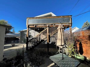 A residential property with a wooden staircase leading to a covered outdoor area, a green trash bin, and a clear blue sky.