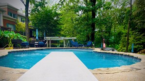 Une piscine dotée d’une terrasse blanche, entourée de verdure et d’arbres.